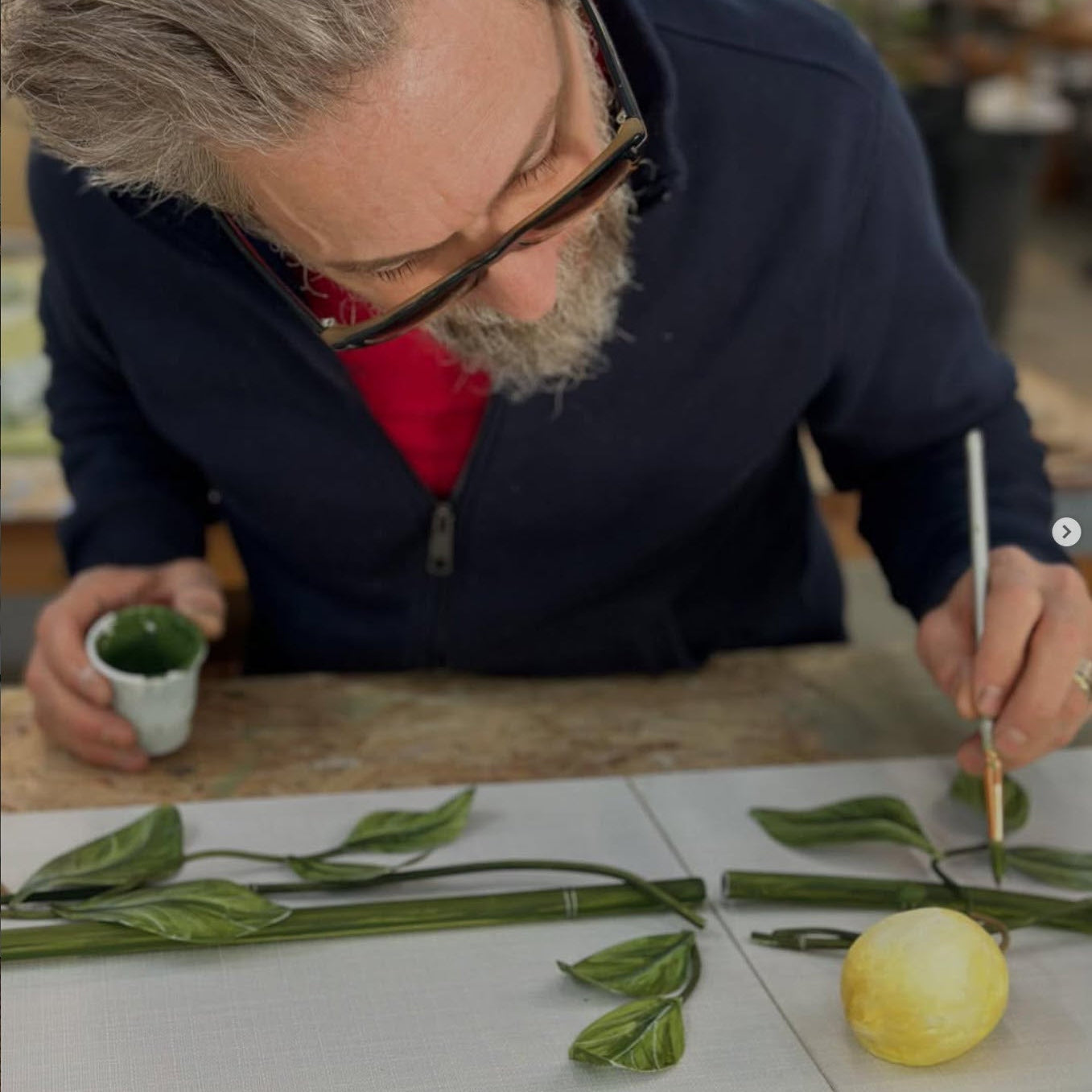 Tommy Mitchell working on a tole floral arrangement with green leaves and a yellow flower on a table.