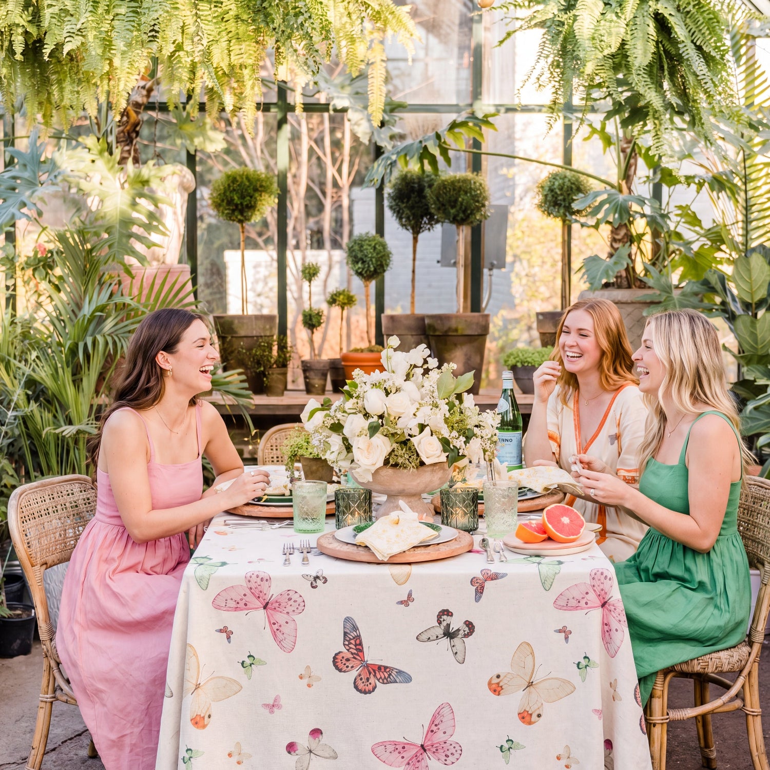 Three women sitting at a table with a butterfly-patterned tablecloth in a plant-filled room.