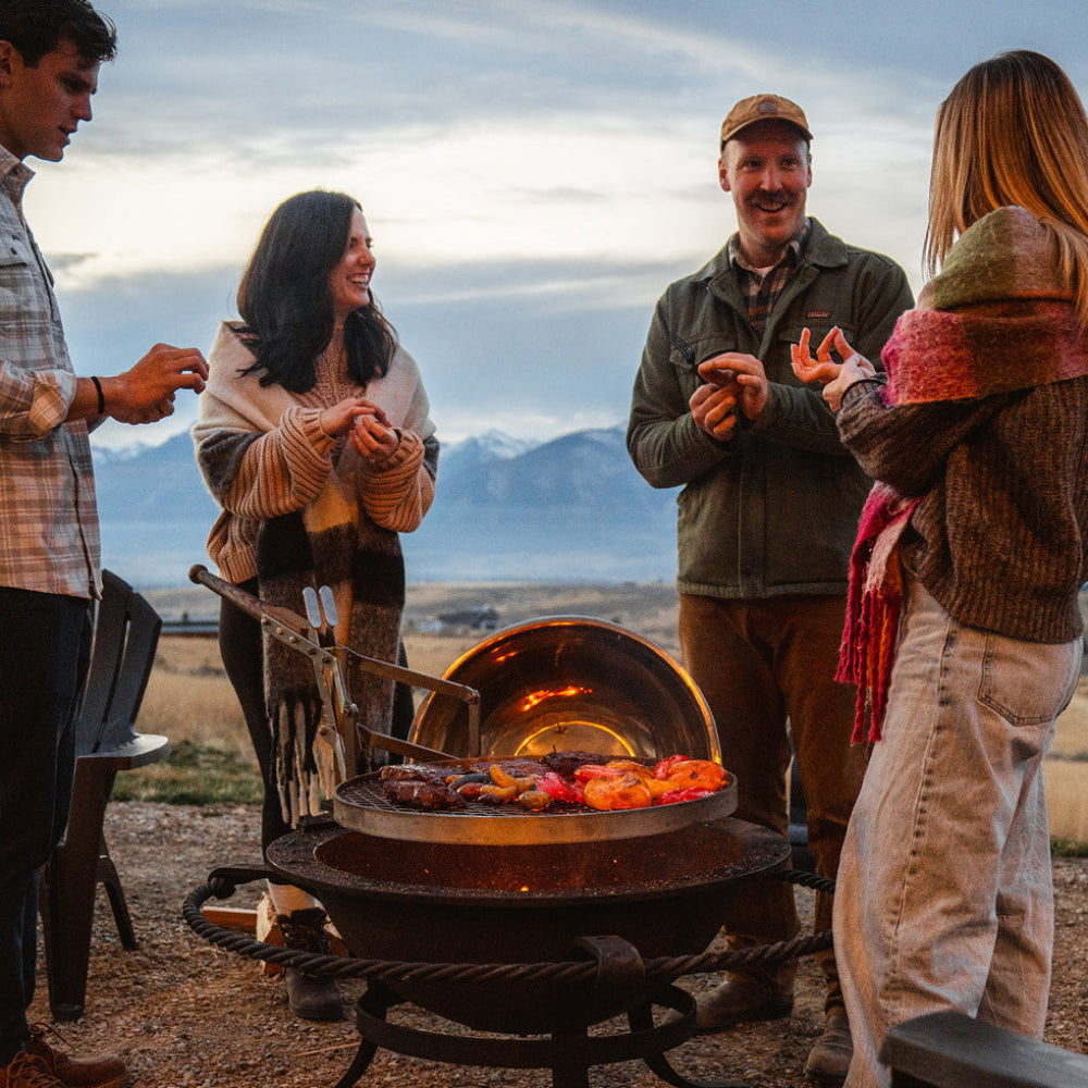 Group of people gathered around a fire pit in an outdoor setting with mountains in the background.