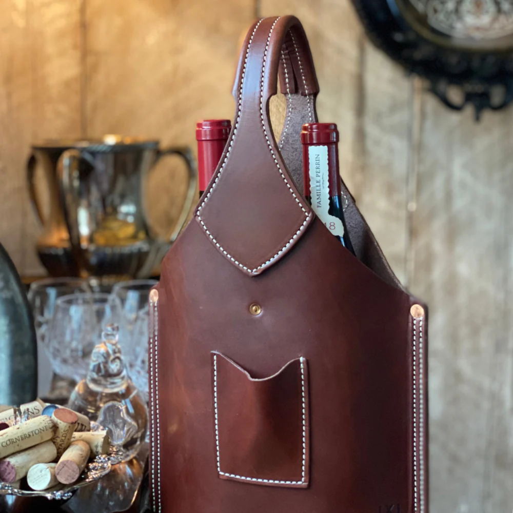 Brown leather wine holder with two bottles inside, on a rustic wooden surface.