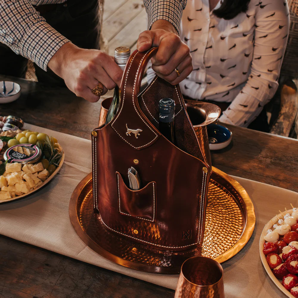 Person holding a brown leather wine tote with a bottle on a wooden table with food.