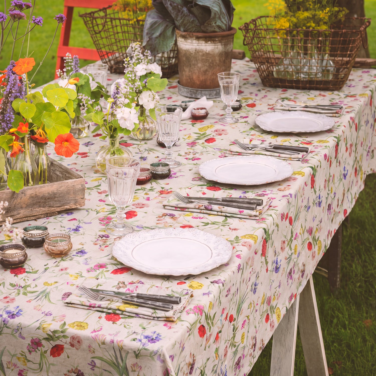 A floral tablecloth spread across an outdoor dining table, styled with white plates, silver cutlery, clear drinking glasses, small metal candle holders, and flower arrangements on a grassy lawn.