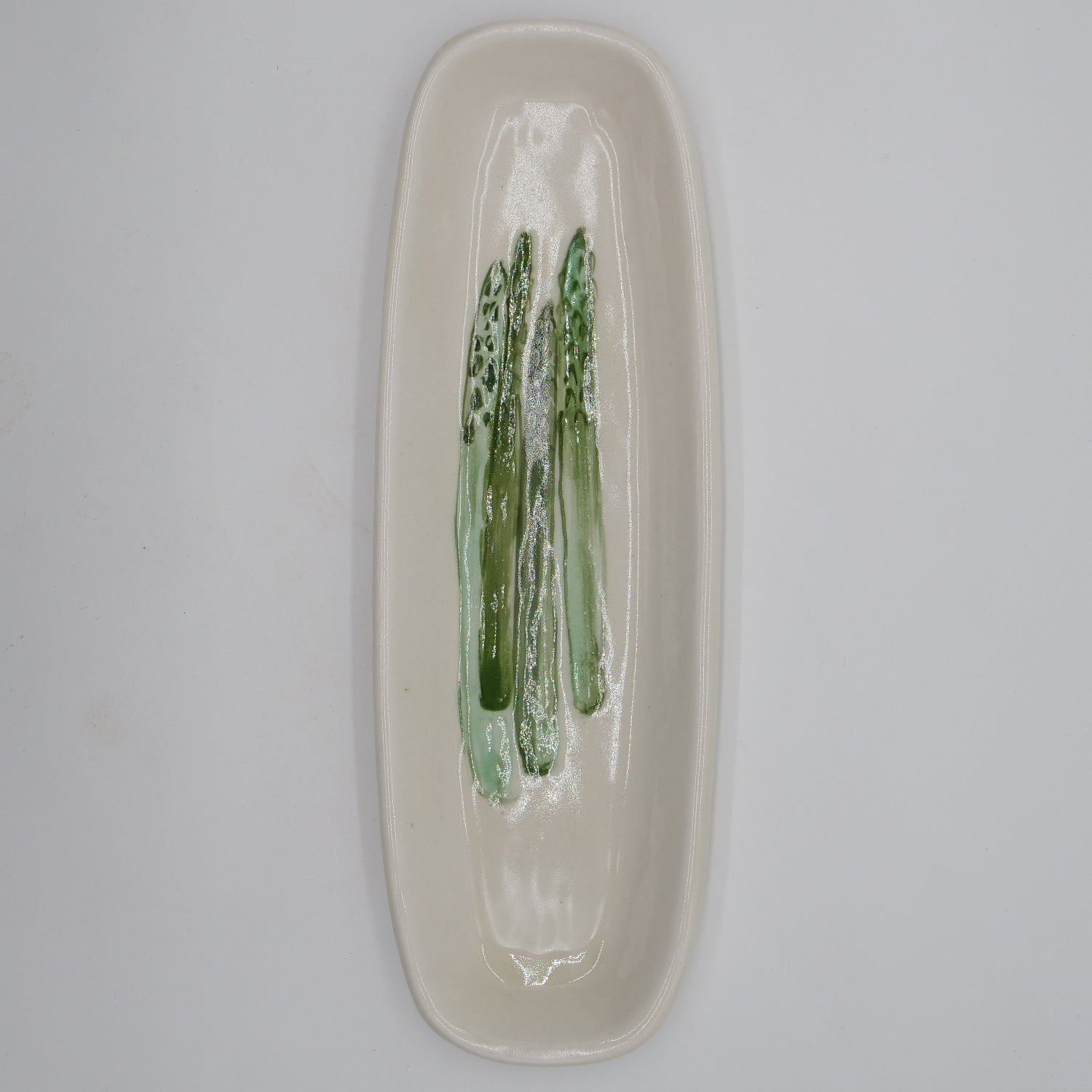 A long, narrow oval ceramic dish with a glossy cream glaze and a painted green asparagus motif in the center, shown on a plain light background.