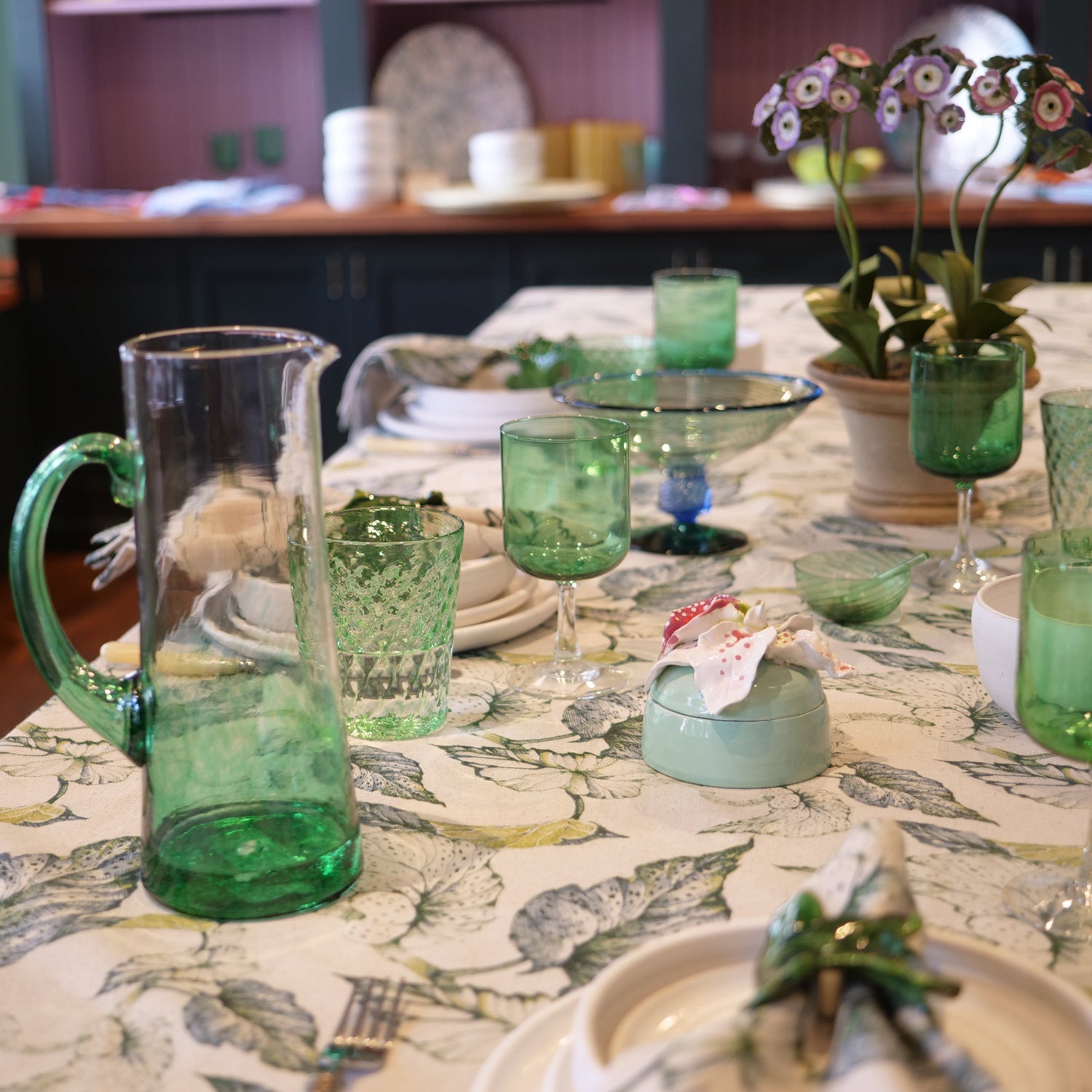 A dining table set with green glassware, including a large green glass pitcher, matching green tumblers, and a green glass bowl, arranged on a linen tablecloth with a botanical caladium leaf pattern.