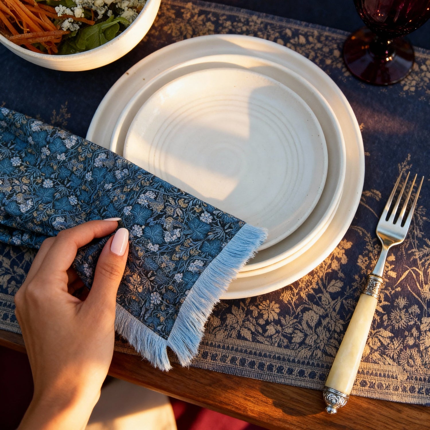 Hand setting a blue Vine Thief patterned napkin on a table with white plates and a fork.